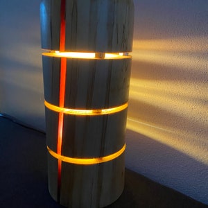 Può includere: Una lampada in legno con una striscia rossa lungo il lato. La lampada è realizzata in legno naturale e ha una luce calda e invitante.