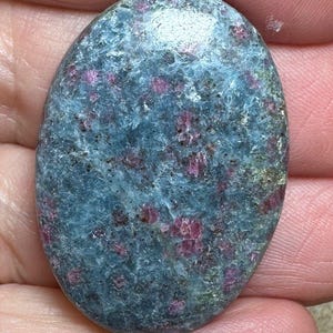 May include: A smooth, oval-shaped cabochon gemstone with a blue-green base and scattered pink inclusions.