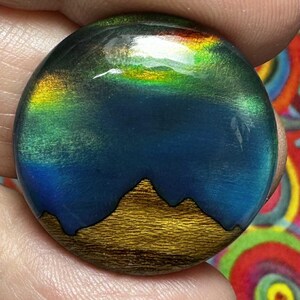 May include: A circular cabochon with an iridescent landscape design. The top has a gradient of green, yellow, and red hues, above a blue area. A gold-coloured mountain range is at the bottom, outlined in black.