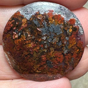 Może przedstawiać: Okrągły, wypolerowany kaboszon agatowy z kolorowym, dendrytycznym wzorem. Kamień ma odcienie czerwonego, pomarańczowego, brązowego, szarego i białego.