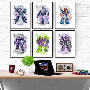 Puede incluir: Seis impresiones enmarcadas de acuarela de personajes de Transformers en varias poses y colores, incluyendo azul, morado, verde y rojo. Las impresiones se exhiben en una pared de ladrillo blanco sobre un escritorio de madera.