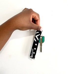May include: Black and white patterned fabric key chain with the word "Positiwax" printed on it. A silver key with a green plastic key cover is attached to the key chain.