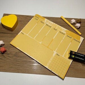 Peut inclure: Un planificateur hebdomadaire jaune avec une grille pour chaque jour de la semaine, y compris lundi, mardi, mercredi, jeudi, vendredi, samedi et dimanche. Le planificateur comprend également une section pour les notes et une grille de calendrier mensuel. Le planificateur est sur une surface en bois avec des formes géométriques jaunes, brunes et roses.