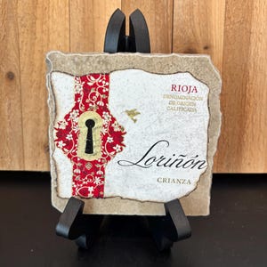 May include: A decorative tile with a keyhole design and the text "Rioja Denominación de Origen Calificada Lorioñon Crianza". The tile is displayed on a black easel against a wooden background.