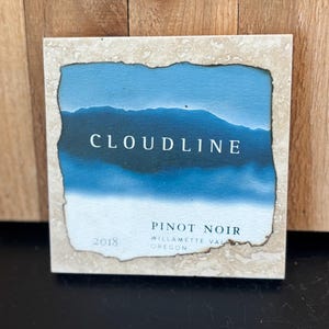 May include: Square coaster with a blue landscape design and the text "CLOUDLINE PINOT NOIR 2018". The coaster has a light beige border with a textured appearance. The background is a wooden surface.