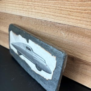 Puede incluir: Una foto en blanco y negro de un coche antiguo está montada en una placa de pizarra. El coche tiene un diseño elegante y futurista con una carrocería redondeada y grandes faros redondos.