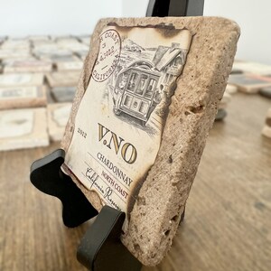 Puede incluir: Un posavasos rectangular de piedra con un soporte negro. El posavasos presenta una etiqueta de estilo vintage con una ilustración de tranvía y el texto "V.NO Chardonnay North Coast California Reserve". El posavasos está hecho de piedra de color marrón claro.