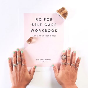 May include: A pink and white workbook with the title "RX for Self Care Workbook" and the text "Love Yourself Daily". The workbook is on a white surface with two hands wearing silver rings and green nail polish.