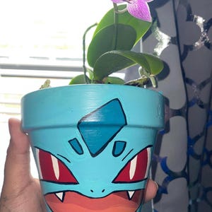 May include: A blue ceramic flower pot painted with a cartoon image of a blue Pokemon with red eyes and a white diamond on its forehead. A pink orchid plant is growing in the pot.
