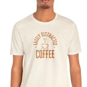 Op de afbeelding: Een wit T-shirt met een bruine afbeelding waarop staat "Easily Distracted by Coffee" met een koffiekopje-icoon.