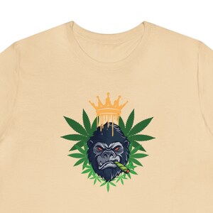 May include: A beige t-shirt with a graphic of a gorilla wearing a gold crown and smoking a marijuana joint surrounded by green marijuana leaves.