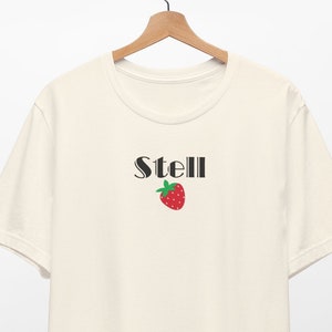 Puede incluir: Una camiseta de color crema con mangas cortas y cuello redondo. La camiseta tiene un gráfico negro de la palabra "Stell" con una fresa roja debajo.