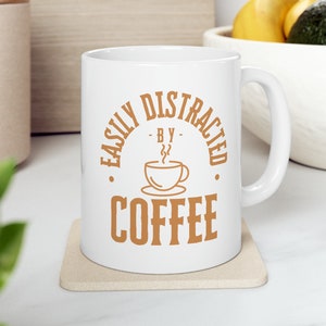 Peut inclure: Une tasse à café en céramique blanche avec un motif brun qui dit "Easily Distracted - By - Coffee" avec une petite icône de tasse à café.