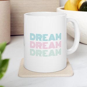 May include: White ceramic mug with a teal, pink, and teal design that says "DREAM" three times. The mug is sitting on a beige coaster.