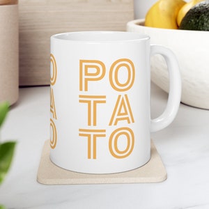 May include: White ceramic mug with a gold outline of the word "POTATO" printed on the front. The mug is sitting on a beige coaster.
