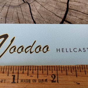 May include: A light blue sticker with black text that reads "Voodoo Hellcaster". The text "Voodoo" is in a stylized script font.