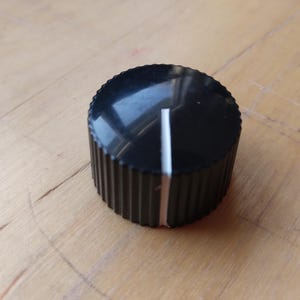 Puede incluir: Un mando de control negro con un borde estriado y una línea indicadora blanca. El mando es cilíndrico y parece estar hecho de plástico. El mando está sobre una superficie de madera.