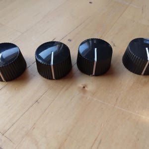 Puede incluir: Cuatro perillas de control negras con una línea indicadora blanca sobre una superficie de madera. Las perillas tienen un borde estriado y una superficie superior brillante, comúnmente utilizadas en equipos de audio.