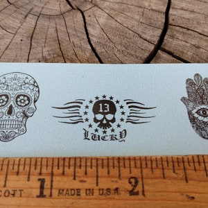 May include: Three black and white tattoo designs on a light blue background. The designs are a sugar skull, a skull with wings and stars, and a hamsa hand with an eye.