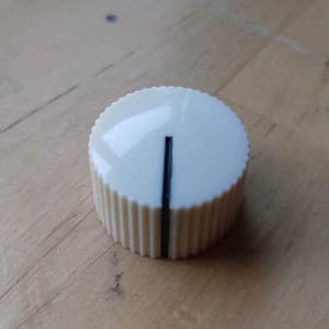 Puede incluir: Un mando de control de color crema con una línea indicadora negra. El mando tiene un borde estriado y una parte superior lisa y redondeada. Este componente se utiliza probablemente para ajustar la configuración de los dispositivos electrónicos.