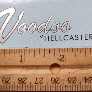 May include: A light blue card with the word "Voodoo" in a stylized silver font, with the text "HELLCASTER" below. A wooden ruler with inch markings is at the bottom of the card, measuring up to 3 inches.
