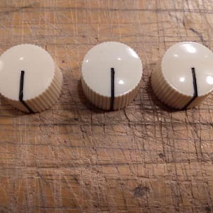Puede incluir: Tres perillas de control color crema con una línea indicadora negra. Las perillas tienen un borde estriado y una parte superior lisa y redondeada. Están dispuestas en fila sobre una superficie de madera, probablemente para un instrumento musical o dispositivo electrónico.