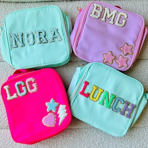May include: Four colorful lunch bags with personalized names and decorative patches. The bags are light blue, pink, green, and hot pink. The names on the bags are NORA, BMG, LGG, and LUNCH. The patches include stars, a heart, a lightning bolt, and a star.
