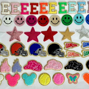 May include: A collection of iron-on patches featuring various designs, including letters, stars, smiley faces, game day pennants, helmets, and more. The patches are all in different colours and styles.