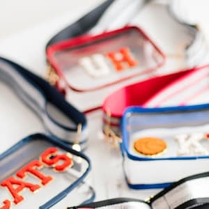 Peut inclure: Quatre sacs à bandoulière en plastique transparent avec des bretelles de couleurs différentes et des lettres brodées. Les sacs sont rouge, bleu et bleu marine. Les lettres sur les sacs sont "UH", "JATS", "BA" et un ballon de basket avec la lettre "K".