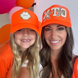 May include: Two people wearing bright orange trucker hats. The hat on the left has a white paw print on it. The hat on the right has the letters "KTC" in white with glitter on it.