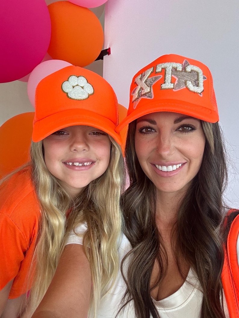May include: Two people wearing bright orange trucker hats. The hat on the left has a white paw print on it. The hat on the right has the letters "KTC" in white with glitter on it.