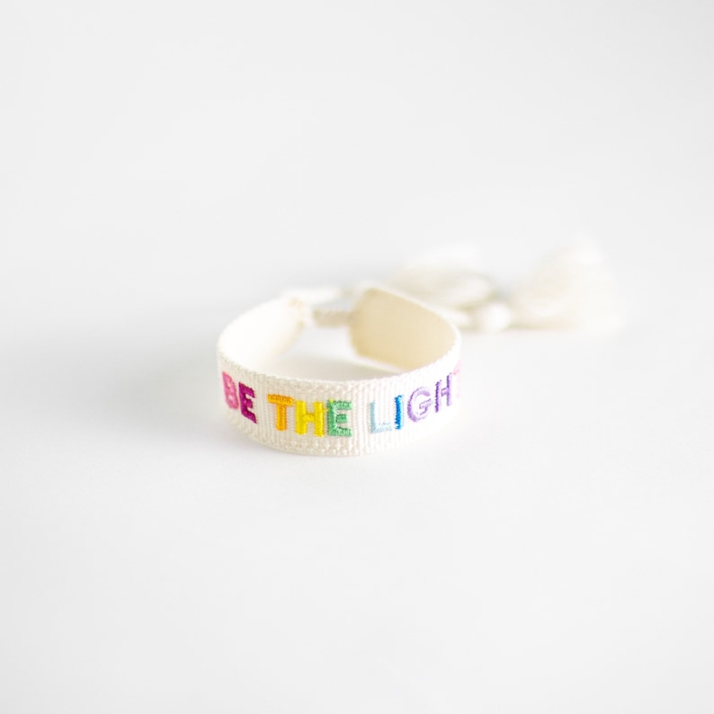 May include: White woven bracelet with a rainbow colored embroidered message that reads "BE THE LIGHT".