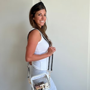 May include: A clear plastic crossbody bag with a black and white striped strap. The bag has a gold chain and is filled with various items, including a gold letter charm that says "T & C".