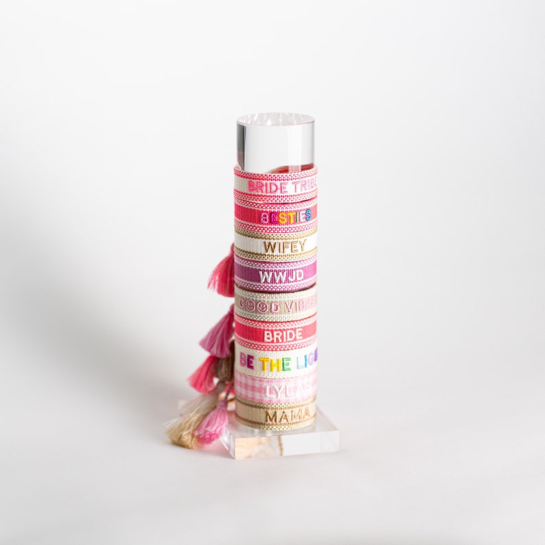 May include: A stack of colorful friendship bracelets with tassels. The bracelets have different phrases on them, including "Bride Tribe", "Besties", "Wifey", "WWJD", "Good Vibes", "Bride", "Be The Light", "Lyla", and "Mama".