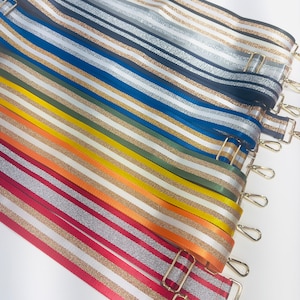 May include: A collection of colorful striped fabric straps with gold glitter accents. The straps have gold metal clasps and are arranged in a stack.
