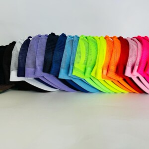 May include: A stack of 20 colourful mesh trucker hats in a rainbow of colours, including black, white, purple, blue, green, yellow, orange, red, pink, and neon pink.