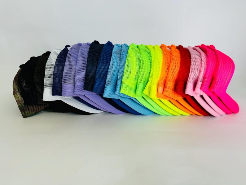 May include: A stack of 20 colourful mesh trucker hats in a rainbow of colours, including black, white, purple, blue, green, yellow, orange, red, pink, and neon pink.
