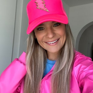 May include: A bright pink trucker hat with a gold lightning bolt design. The hat is made of mesh and has a curved brim.