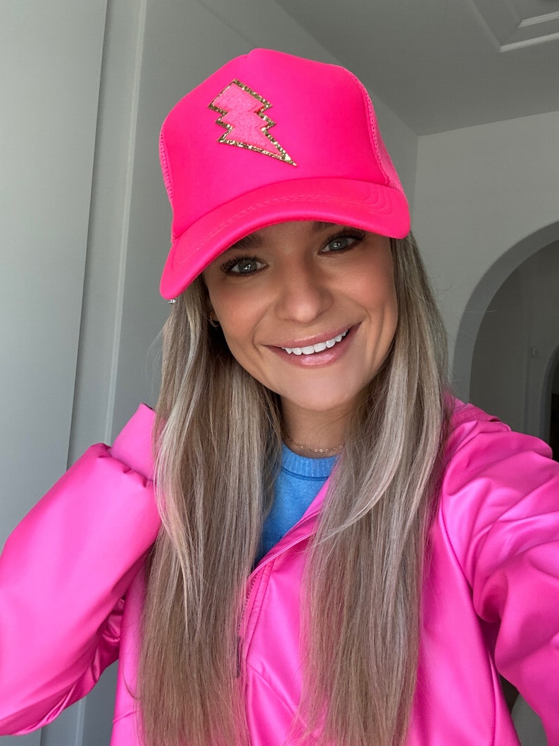 May include: A bright pink trucker hat with a gold lightning bolt design. The hat is made of mesh and has a curved brim.
