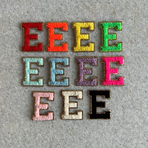 May include: Ten felt letter patches in various colours, each featuring the letter "E" in a gold outline. The colours include red, orange, yellow, green, light blue, dark blue, purple, pink, white, and black.