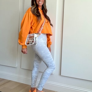 May include: A woman wearing a bright orange cropped hoodie, white camouflage leggings, and orange sneakers. She is carrying a clear crossbody bag with a brown strap and the initials "TC" on the front.