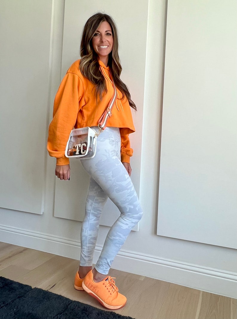 May include: A woman wearing a bright orange cropped hoodie, white camouflage leggings, and orange sneakers. She is carrying a clear crossbody bag with a brown strap and the initials "TC" on the front.