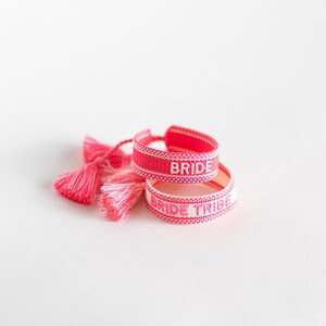 May include: Two pink and white woven friendship bracelets with tassels. The top bracelet says "BRIDE" and the bottom bracelet says "BRIDE TRIBE".