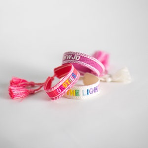 May include: Three colorful friendship bracelets with tassels. The bracelets are pink, white, and red with rainbow lettering. The lettering on the bracelets reads "WWJD", "Besties", and "The Light".