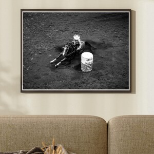 May include: Black and white photograph of a rodeo scene. A rider on horseback is in motion near a barrel. The image is framed in a black border and mounted on a wall. A sofa is in the foreground.