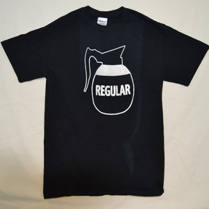 Op de afbeelding: Zwart T-shirt met een witte afbeelding van een koffiekan met het woord "REGULAR" erop gedrukt.