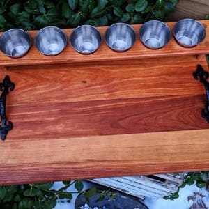 May include: A wooden serving tray with black metal handles. The tray has a built-in rack with six stainless steel cups.
