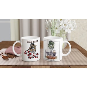 Op de afbeelding: Twee witte keramische mokken met humoristische skeletontwerpen. Eén mok leest "DEAD INSIDE" met een skelet met een zonnebril en een bloemenhoofdband. De andere mok zegt "BUT CAFFEINATED" met een skelet dat een koffiekopje vasthoudt.