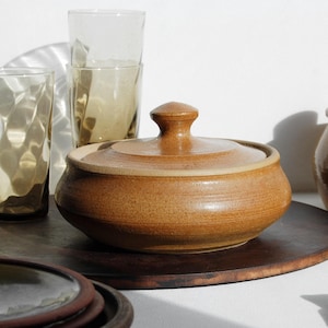 May include: A selection of handmade pottery and glassware. The image shows a lidded serving bowl, jars with lids, and textured drinking glasses. The pottery has a warm, earthy tone, and the glasses have a subtle amber hue. A wooden tray and plates are also visible.