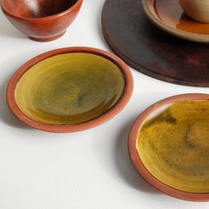 May include: An overhead shot featuring several ceramic items. Two small, oval plates with a yellow-green interior and a reddish-brown rim are in the foreground. A small bowl and a larger plate are also visible.